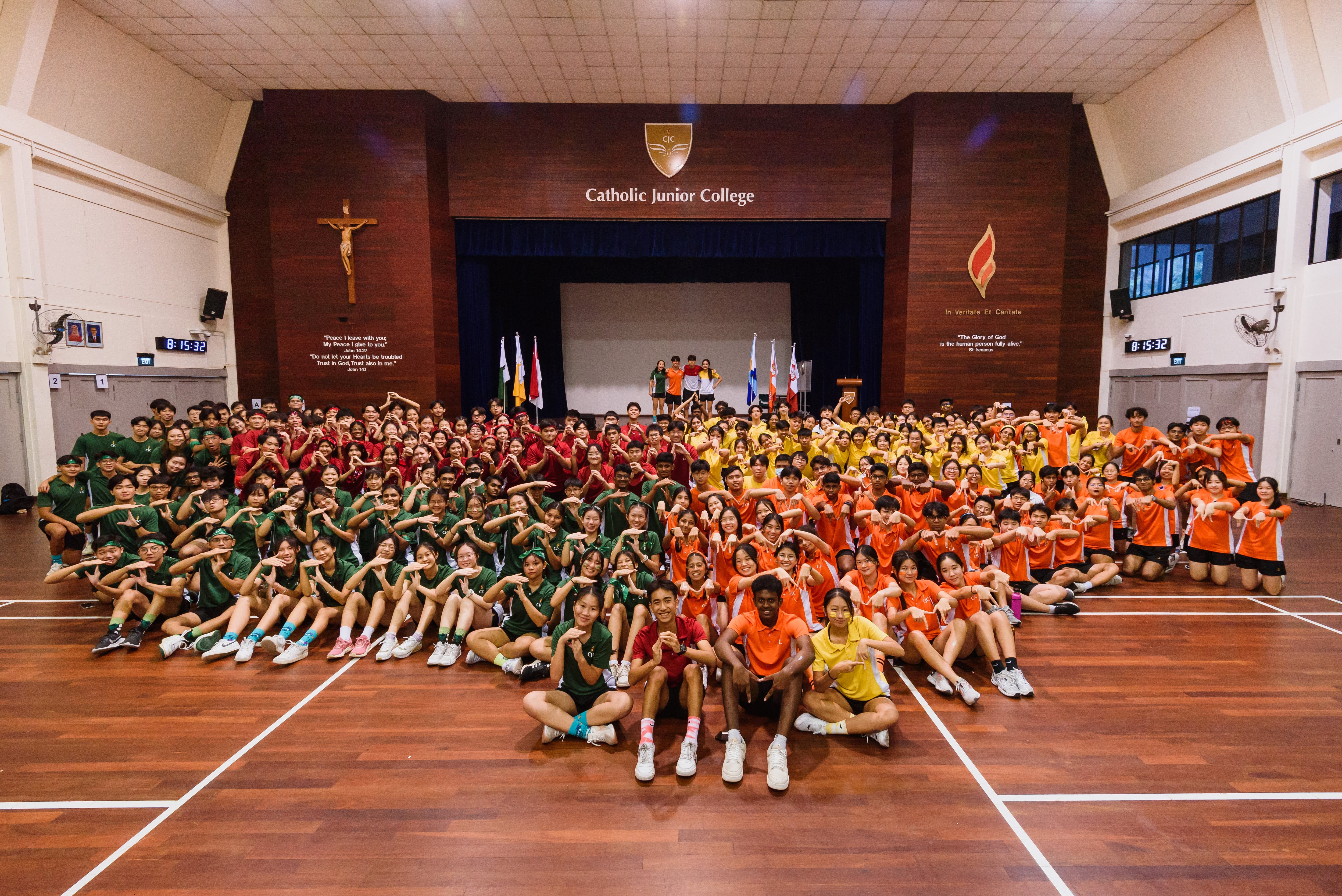 Large group of students in green, red, yellow, and orange shirts seated in a school hall, posing.